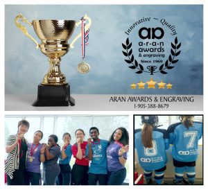 "A collage featuring a golden trophy and medal on a blue background adorned with Aran Awards & Engraving's logo, name, number, and five gold stars. Below, a group of seven office workers proudly display their winning medals supplied by Aran Awards & Engraving. Another image shows two female minor hockey players in full dress, backs turned to the photographer, watching the game from behind the glass, their jerseys proudly displaying Aran Awards & Engraving's logo. Aran Awards & Engraving proudly supports their team.